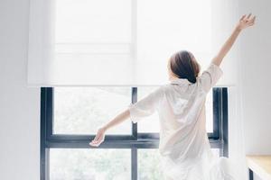 espalda ver de joven mujer elevado su manos y extensión su cuerpo por poco el ventana después su despertar arriba en el Mañana. conceptual de mujer estilo de vida. foto