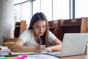 joven asiático estudiantes en casual vestir hacer deberes y utilizar un tecnología ordenador portátil en el Universidad biblioteca. el estudiantes son buscando para conocimiento y son preparando para el examen. foto