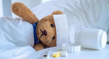 The teddy bear and the fever-reducing patch lie on the bed in a white-tone bed next to a medicine table and body temperature monitor. Used for health insurance or illness concepts photo