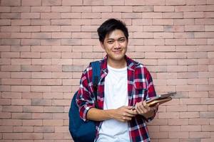 asiático masculino estudiantes vestir tartán camisas en pie siguiente a un ladrillo muro, que lleva un mochila, que lleva libros, colegio suministros, preparando para estudiar, sonriente. foto
