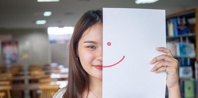 un hermosa hembra estudiante en Asia sostiene un papel dibujo un sonrisa con rojo tinta y un contento postura en instalaciones. foto