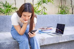 un asiático hembra estudiante es sentado en el jardín siguiente a el teléfono, mirando a el resultados de un en línea prueba en un teléfono inteligente, en el cara de decepción y lamentar terminado el información en el teléfono foto