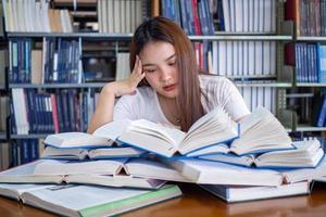 hembra estudiantes son estresado con leyendo un lote de libros metido en el mesas en el biblioteca. a preparar para el examen foto