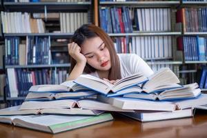 hembra estudiantes son estresado con leyendo un lote de libros metido en el mesas en el biblioteca. a preparar para el examen foto