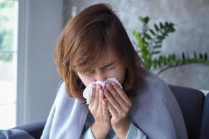 asiático mujer tener líquido nariz y captura un frío, tos, estornudos, fiebre, sentado enfermo en el sofá dentro el casa. foto