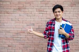 asiático masculino estudiantes vestir tartán camisas en pie siguiente a un ladrillo muro, que lleva libros, colegio suministros, preparando para estudiar. foto