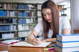 asiático hembra estudiante sentado a un escritorio y escritura en un cuaderno. joven hembra estudiantes hacer deberes en el biblioteca foto