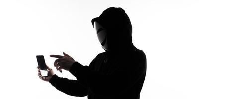 hacker anónimo y mascarilla con smartphone en la mano. hombre con camisa de capucha negra sosteniendo y usando un teléfono móvil con fondo blanco. representan el concepto de piratería informática o robo de datos personales foto