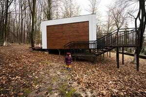 Baby girl against one-storey modular house in spring rainy forest. photo