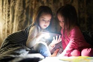 Two sisters watching movie or cartoon in smartphone together with blanket cover in bed at night home. photo