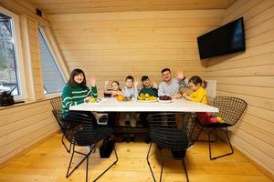 Family with four kids eat fruits in wooden country house on weekend. They wave hands at the camera. photo