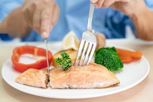 Asian senior woman patient eating Salmon steak breakfast with vegetable healthy food while sitting and hungry on bed in hospital. photo