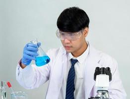 estudiante científico asiático en un laboratorio de mezcla de reactivos en un laboratorio de investigación científica con tubos de ensayo de varios tamaños y microscopios. sobre la mesa en el fondo blanco de laboratorio de química de laboratorio. foto