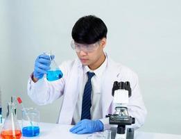 estudiante científico asiático en un laboratorio de mezcla de reactivos en un laboratorio de investigación científica con tubos de ensayo de varios tamaños y microscopios. sobre la mesa en el fondo blanco de laboratorio de química de laboratorio. foto