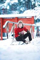 Smiling young cheerful caucasian man is wearing worm red sweatshirt and fun winter ear hat is having fun with sled in a snowy park or forest on winter day. Happy Christmas winter holiday photo