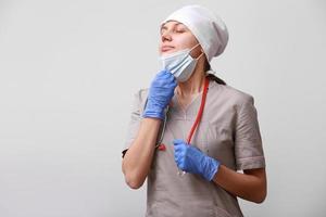 sonriente hembra médico con estetoscopio en uniforme. mujer es tomando apagado médico protector máscara desde cara a respirar aire, en blanco antecedentes. dificultades y problemas a trabajo de un médico obrero. foto