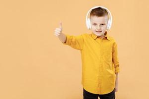 Portrait of funny clever school boy with headphones in yellow shirt. Yellow studio background. Education. Looking, smiling and shows a thumb up at camera. High quality photo