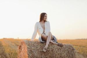 Young woman with long hair, wearing jeans skirt, light shirt and straw bag in hand, sitting on bale on field in summer. Female portrait in natural rural scene. Environmental eco tourism concept. photo