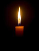 A single burning candle flame or light glowing on an orange candle on black or dark background on table in church for Christmas, funeral or memorial service with copy space photo