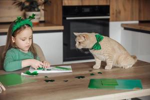 un pequeño niña con un vendaje en su cabeza sorteos y cortes verde tréboles para S t. patrick's día a un mesa a hogar en el cocina, siguiente a su es su hermosa gato con un verde arco Corbata alrededor su cuello foto