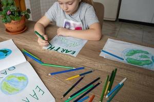 A child at home at the table draws the planet Earth with a world map with multi-colored pencils and felt-tip pens on white paper. Peace and earth day concept. photo