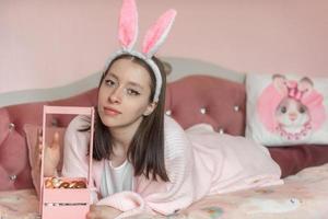 A beautiful young girl with hare ears on her head lies on the bed at home with a basket of decorated Easter eggs. Preparation for Easter. photo