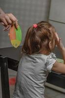 The older sister glues a paper fish to her little sister on her back at home in the kitchen, the child has breakfast. Funny family jokes, April Fool's Day celebration photo