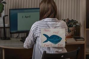 A young woman with a paper fish from behind indoors works at a computer on which the inscription the first of April, April Fool's Day, jokes and jokes in the office on top of each other photo