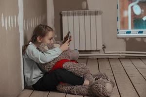 a sad nine-year-old girl, she is sitting in a room in the attic without repairs on the floor, she hugged an old big teddy bear, she has a phone in her hands, she communicates on social networks photo