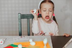 a little girl decorates white eggs with stickers with different emotions, in her hands is a white egg with emotions, the child copies and shows with facial expressions. photo