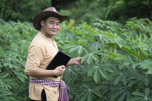 Handsome Asian man farmer is at cassava garden, holds smart tablet to check quality of growth and plants disease. Plan agricultural market online. Concept , Smart farmer. Technology agriculture. photo