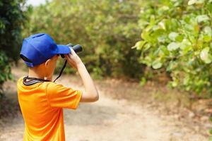 Asian boy wears blue cap, orange shirt, holds binocular in forest. Concept, Exploration. Adventure activity. Learning by doing. Summer camp. Real life experience. Nature survey. photo