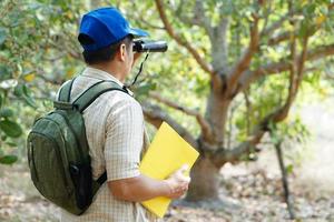 Asian man explorer wears blue cap, holds binocular in forest to survey botanical plants and creatures wildlife. Concept, nature exploration. Ecology and Environment. photo
