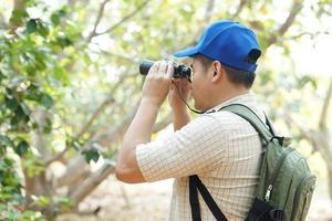 Asian man explorer wears blue cap, holds binocular in forest to survey botanical plants and creatures wildlife. Concept, nature exploration. Ecology and Environment. photo
