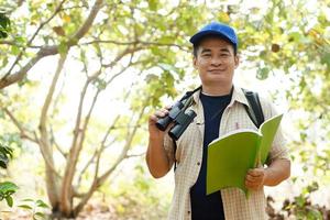 asiático hombre explorador usa azul gorra, sostiene binocular en bosque a encuesta botánico plantas y criaturas fauna silvestre. concepto, naturaleza exploración. ecología y ambiente. foto