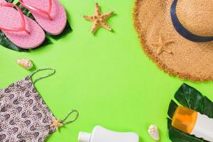 Summer accessories with t-shirt, seashells, flip flops sunscreen bottle and straw hat on green background top view flat lay