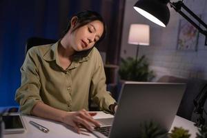 Asian woman talking on smartphone and working on desktop pc late at home at night, can not sleep photo