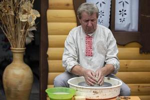 Belarus, the village of Lyaskovichi. August 20, 2022. A holiday of ethnic cultures. An elderly Slavic man in a Belarusian linen embroidered shirt sculpts items from clay. Ethnic crafts. photo