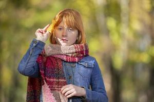 Red-haired little girl wrapped in a woolen scarf with a yellow maple leaf. Child in the autumn park.The concept of autumn has come. photo