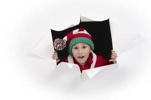 A funny child in a Santa Claus hat climbs out of paper. photo