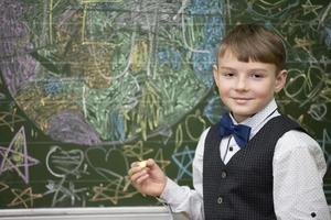 A handsome boy of school age stands at the blackboard, looks at the camera and smiles. Smart student. Back to school. photo