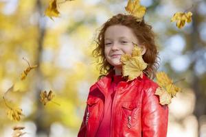 contento pequeño niño, Pelirrojo niña se ríe y obras de teatro con otoño hojas en naturaleza al aire libre foto