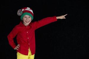 contento chico en Papa Noel claus sombrero posando en negro antecedentes. un niño en nuevo años ropa puntos un dedo a un vacío lugar. foto