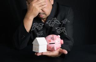 paper house and piggy bank on hand on black background, Businessman stressed about money to buy a new house, saving money concept photo