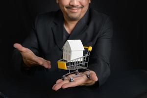 Paper house in a shopping trolley on a salesman hand on black background. photo