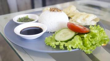A plate containing of a white rice, lettuce, a slice of tomato and cucumber, crackers, soy sauce and crushed green chilies photo