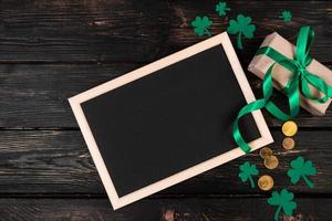 Empty frame for text, gold coins, clover shamrock and gift with green ribbon on a dark wooden background. Good luck symbols for St. Patrick's Day. photo