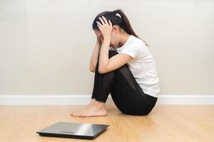 Depressed and stress, sad asian young woman,girl cover face with hand, sitting on floor, weighing on scale in front of her. Gain lose weight and overweight fat. People diet, unhealthy, unhappy concept photo