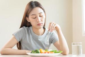 Diet in bored face, unhappy beautiful asian young woman, girl on dieting, holding fork in salad plate, dislike or tired with eat fresh vegetables. Nutrition of clean, healthy food good taste. photo