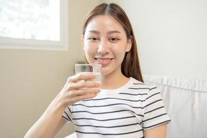Happy beautiful, asian young woman, girl drinking, sip fresh glass of water for hydration of body, holding transparent glass in her hand, thirsty at home. Health care, healthy lifestyle concept.